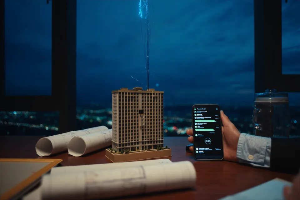 A building model on a desk at dusk, a thread of light rising from it, a hand holding a phone beside it